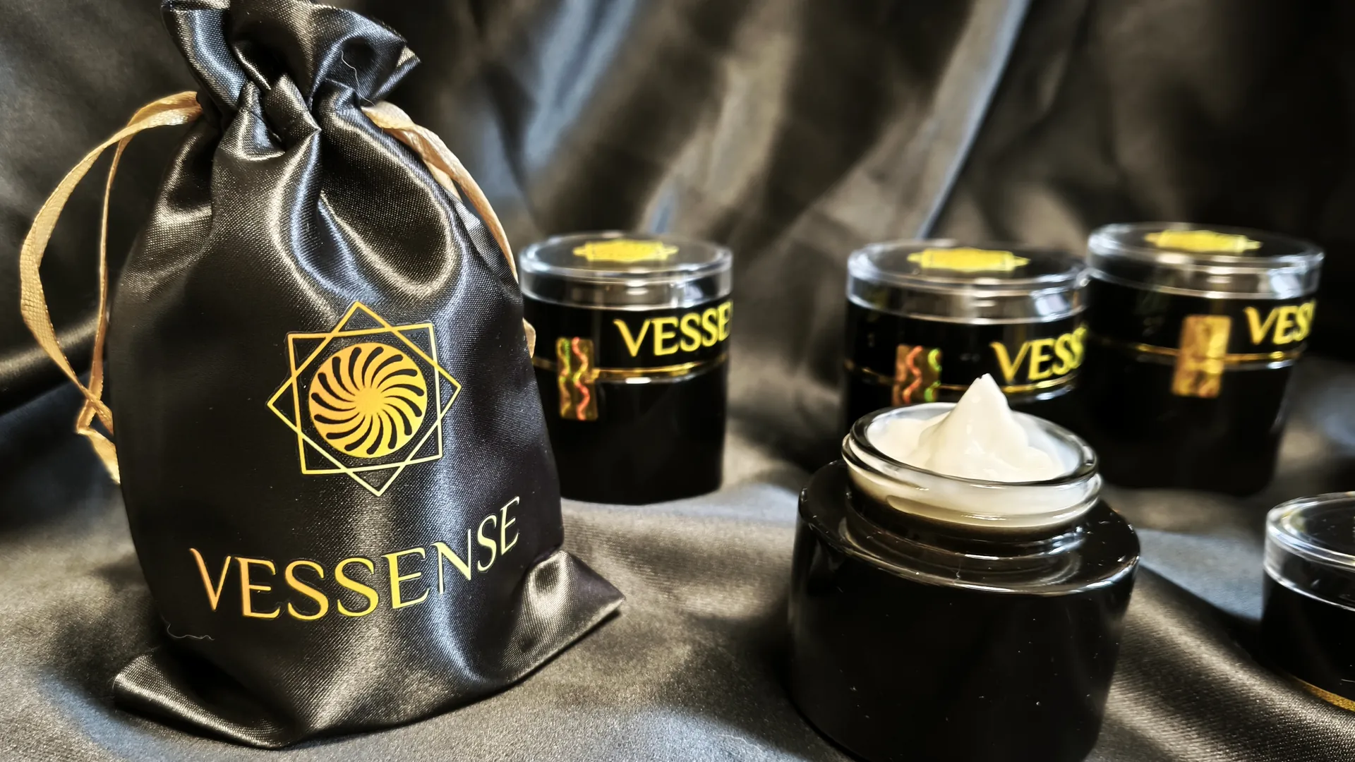 Collection VESSENSE sur satin noir — pochette de satin, pots scellés et pot de crème ouvert